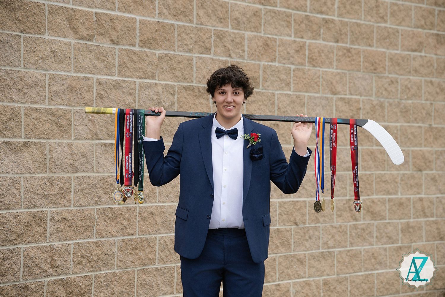 Graduate in formal attire displaying athletic medals against brick wall background.