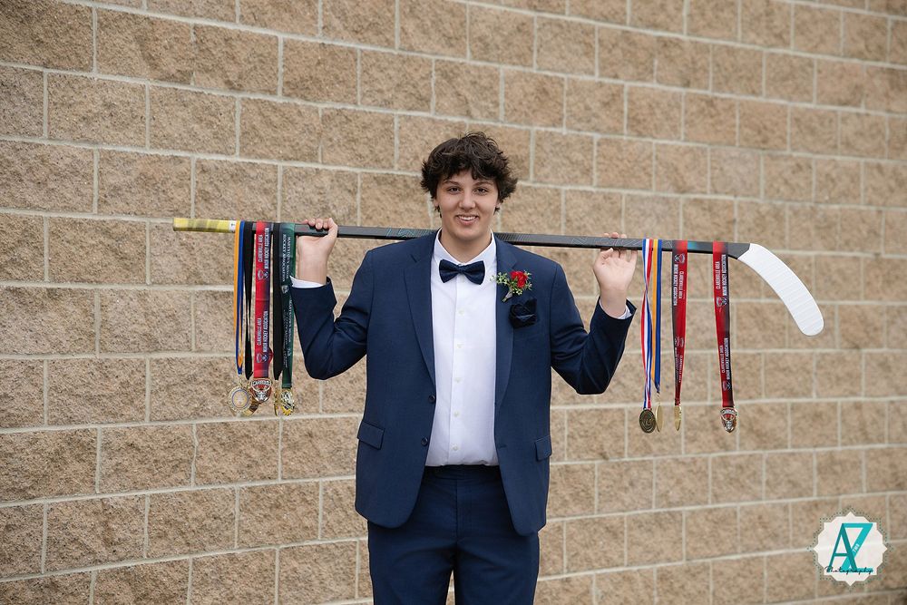 Graduate in formal attire displaying athletic medals against brick wall background.
