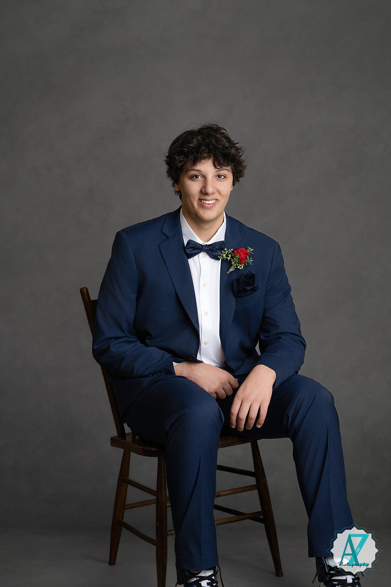 A formal portrait shows a person in a navy suit and bowtie seated on a wooden chair against a gray backdrop.