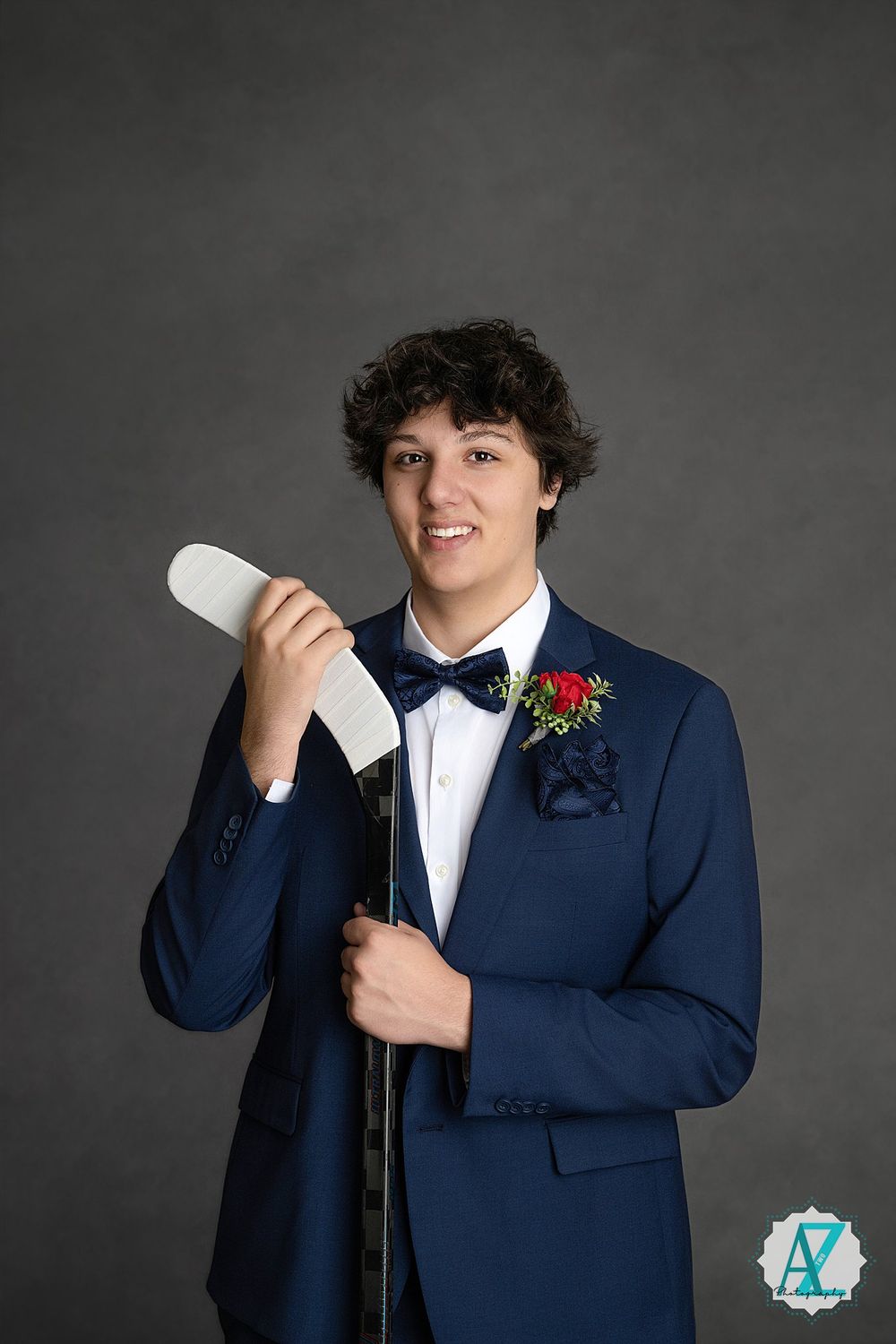 Student in navy blue suit and bow tie poses with diploma for formal portrait against dark gray background.