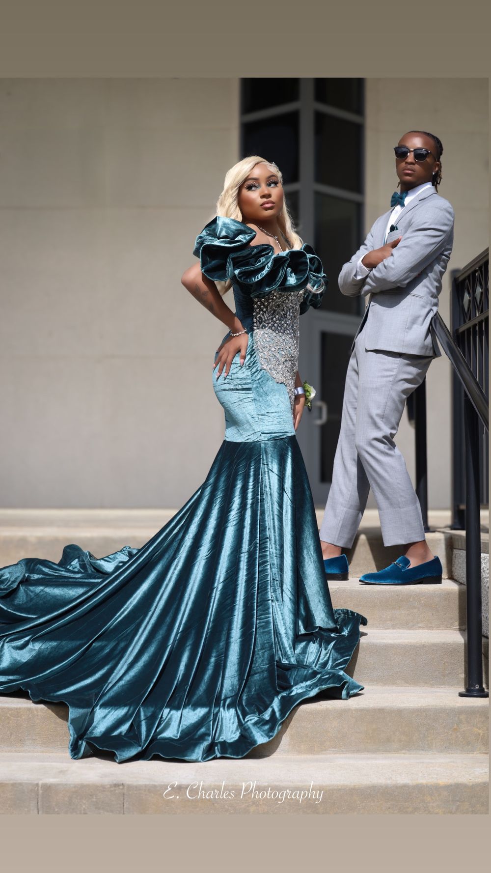 Stunning teal mermaid prom gown with ruffled shoulders on steps, escort in gray suit behind her.