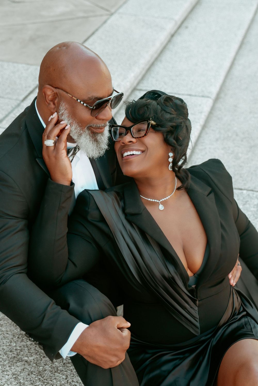 Joyful Black couple in black formal wear sitting on steps, sharing a laugh and tender moment.