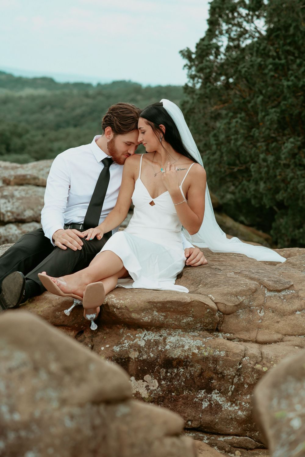 Bride and groom sit closely on rocky outcrop, foreheads touching, surrounded by lush green forest backdrop.