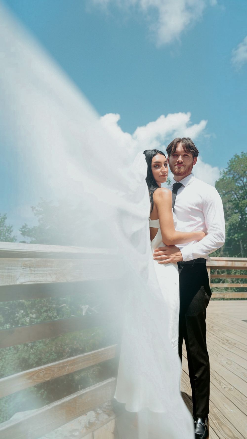 A bride in a flowing white gown and veil embraces a groom in a black suit on a sunny outdoor deck.