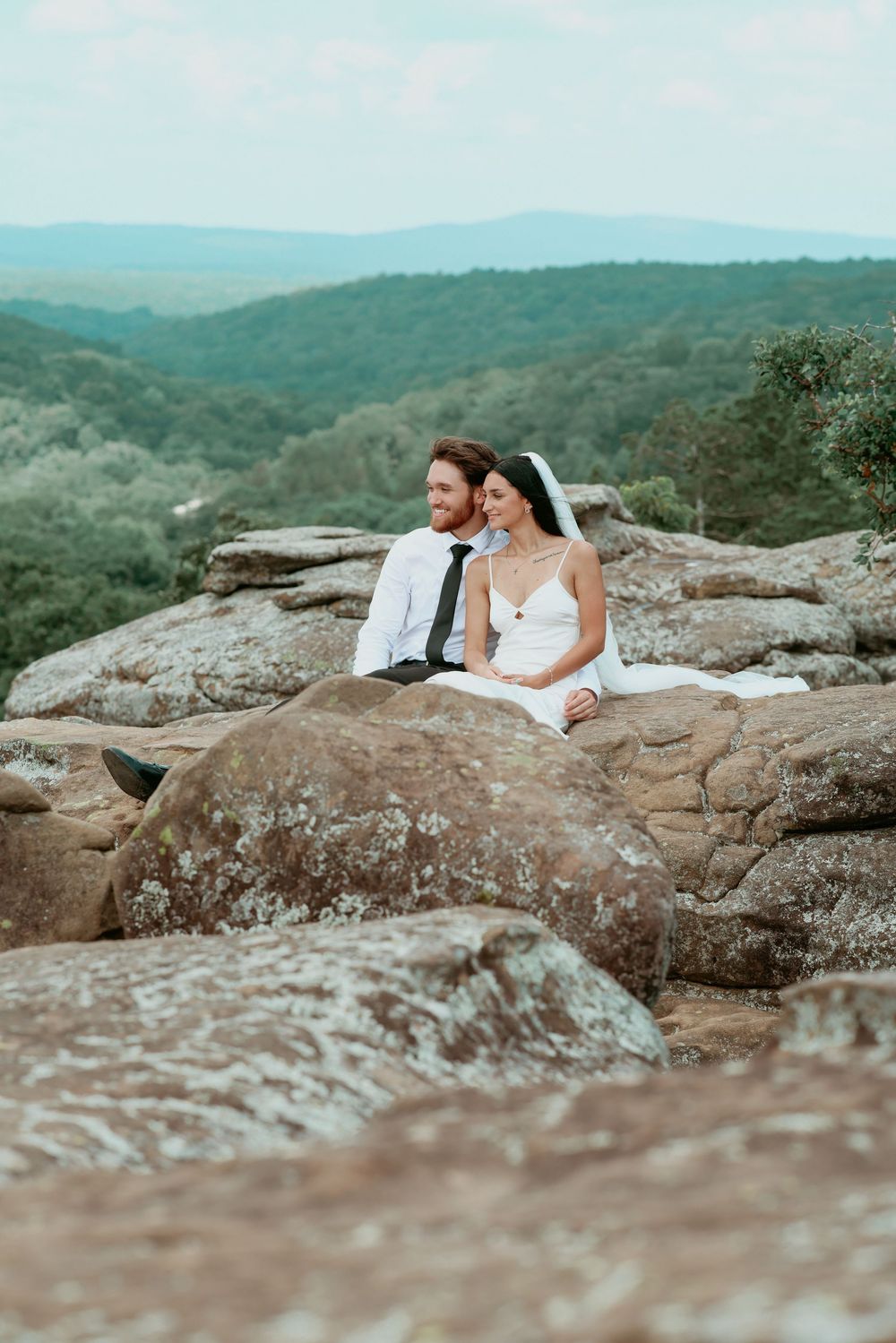 Bride and groom sitting on rocky overlook with lush green mountain landscape behind them on their wedding day.