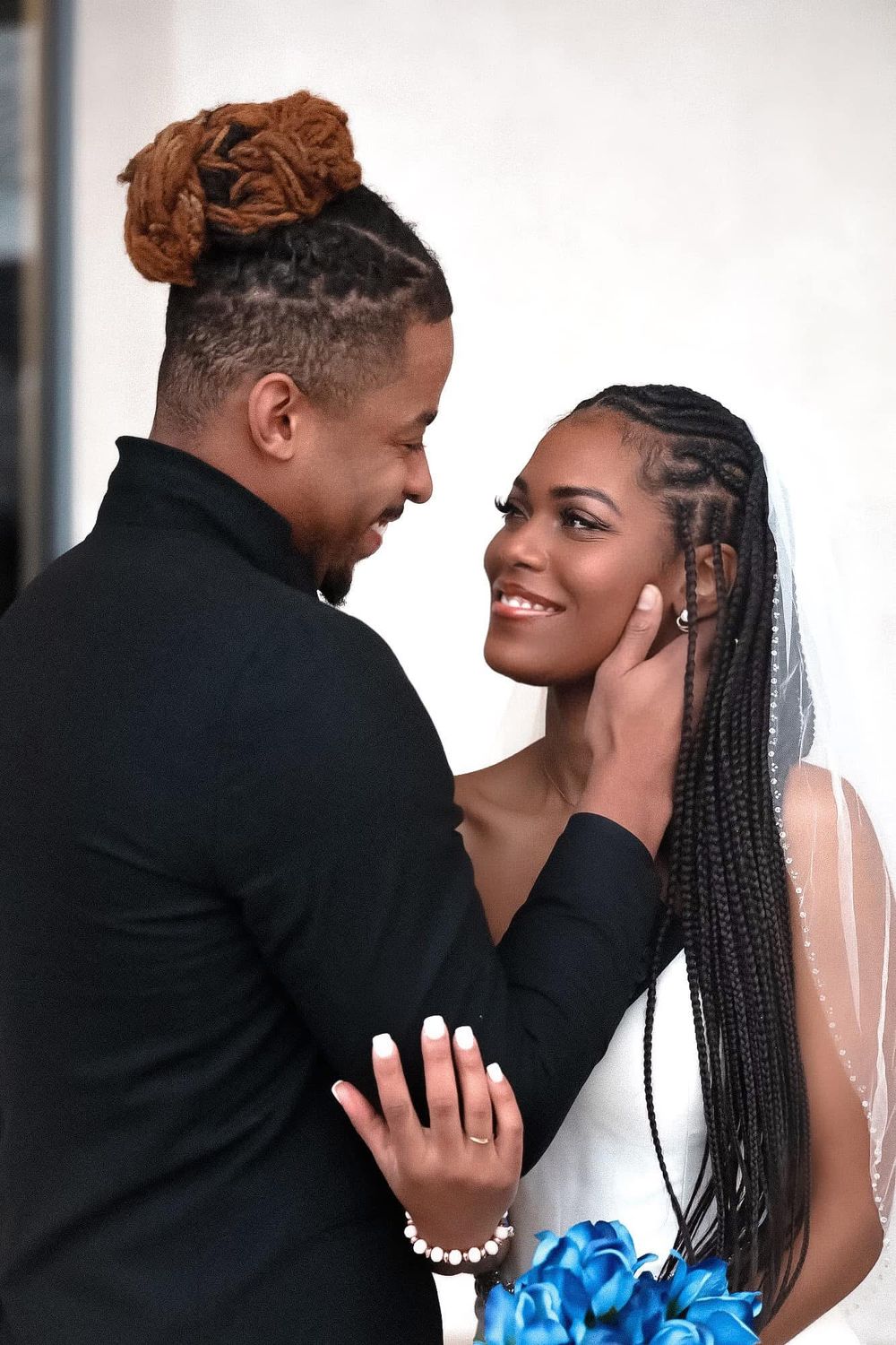 A groom tenderly cups the face of his smiling bride in braids and veil, holding blue flowers, in wedding attire.