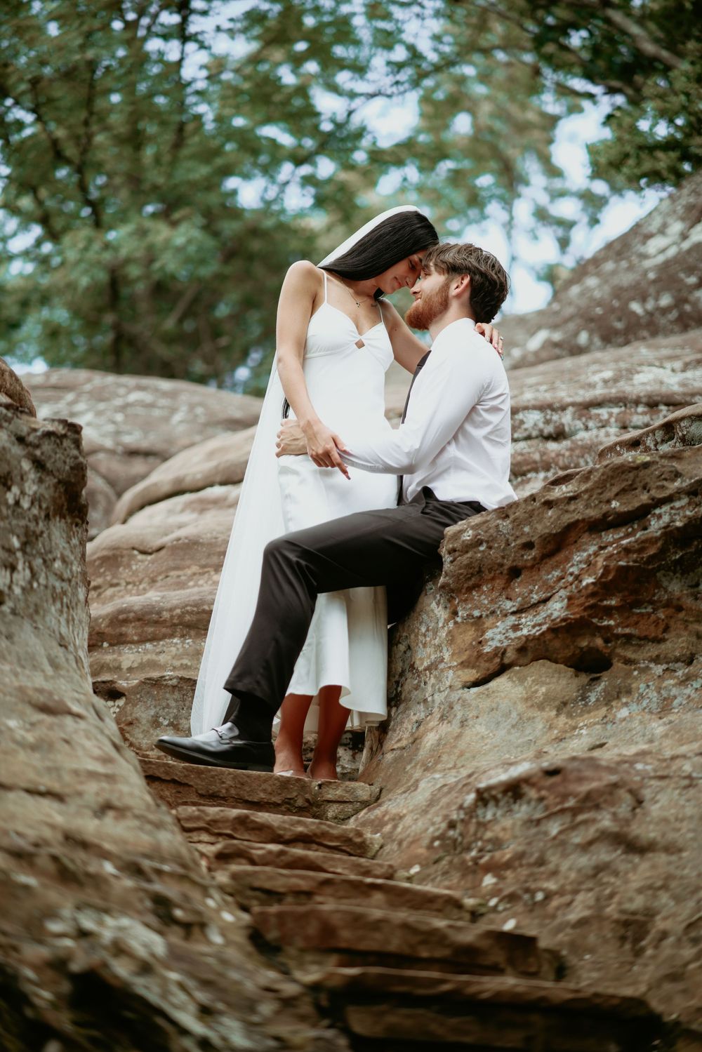 A bride and groom share a romantic kiss while sitting on rocky terrain surrounded by lush green trees.