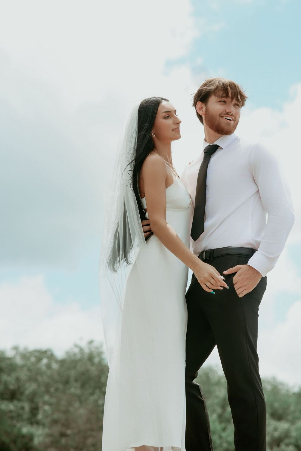 Bride in white gown and veil stands with groom in black suit and tie outdoors under a cloudy sky.