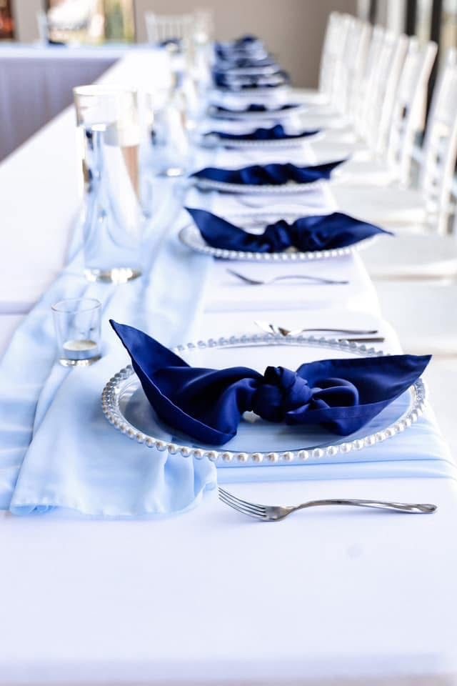 Elegant wedding reception table set with navy blue napkins on silver charger plates with white linens.