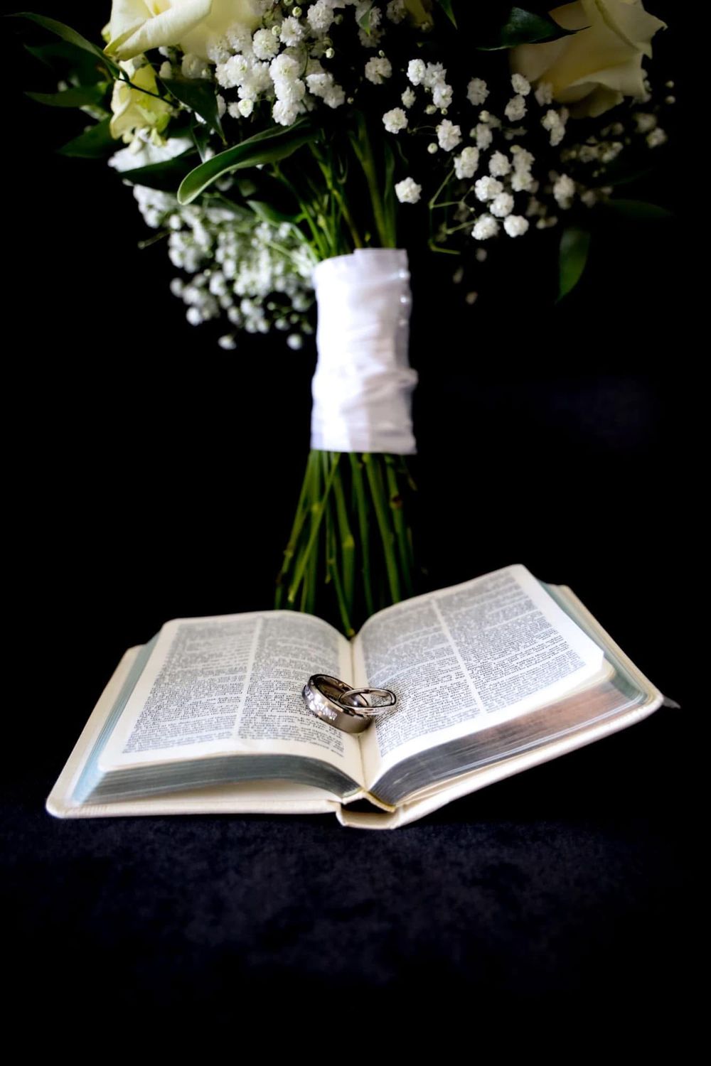 Wedding rings resting on open Bible with white rose bouquet and baby's breath on dark background.