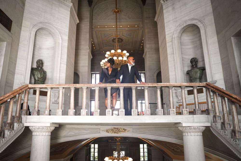 Couple standing on a grand interior balcony with chandelier and classical architecture.