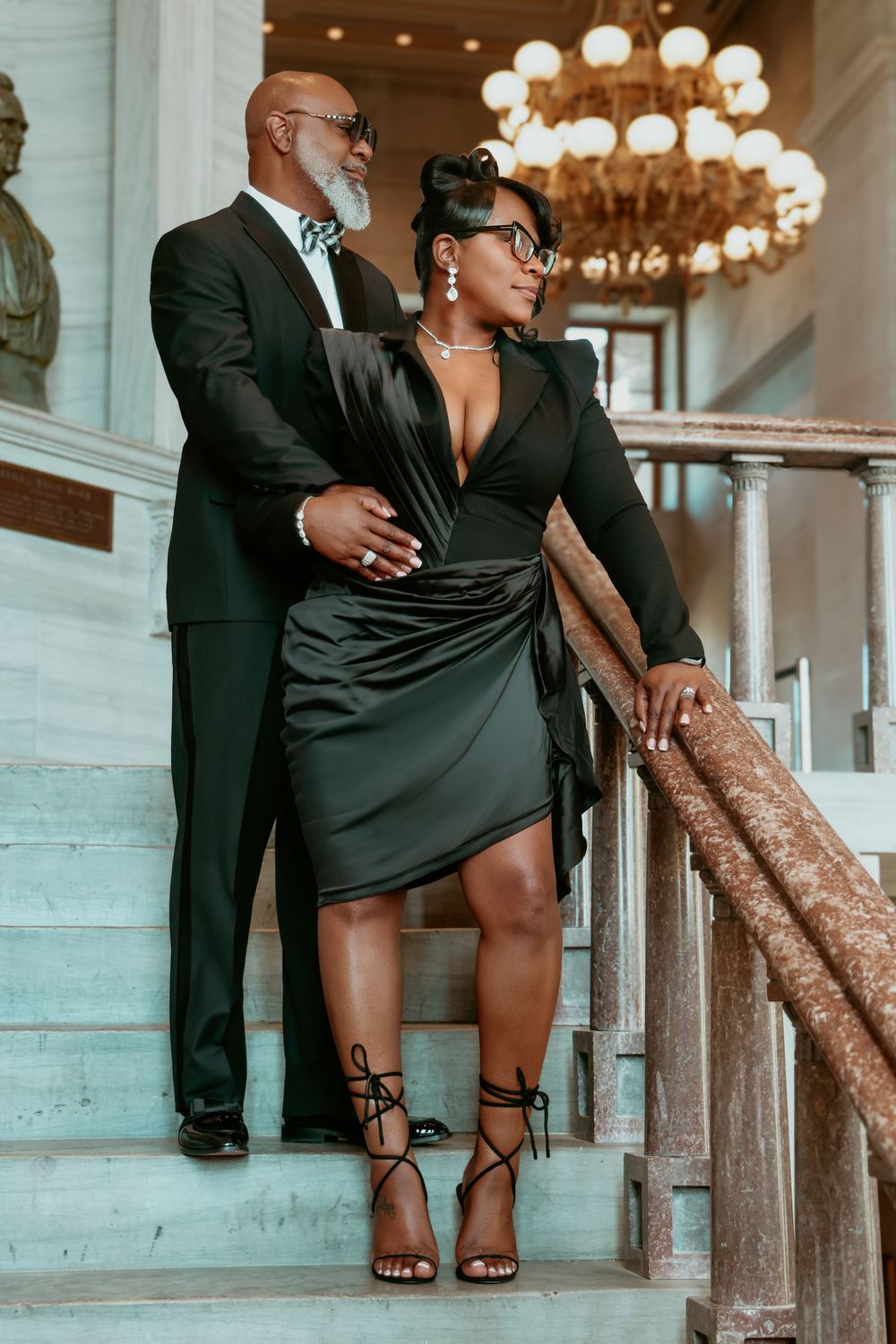 Sophisticated Black couple posing on marble stairs under a glowing chandelier in black formal wear.