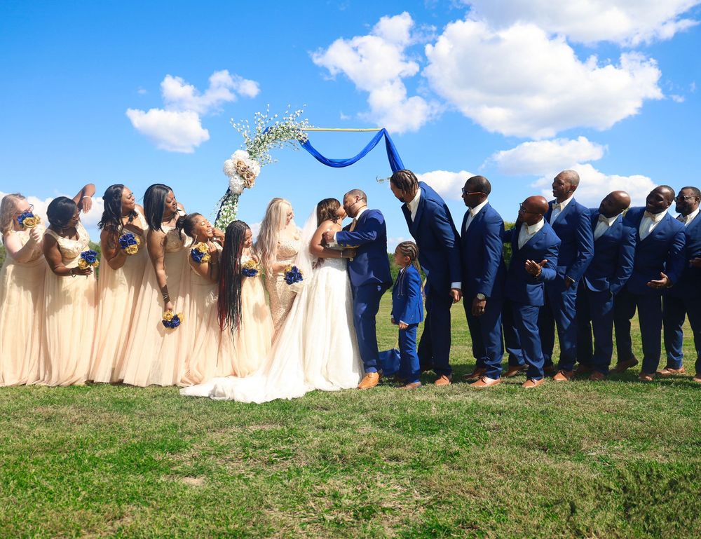 Wedding party poses under floral arch, bridesmaids in champagne gowns and groomsmen in navy suits on sunny day.