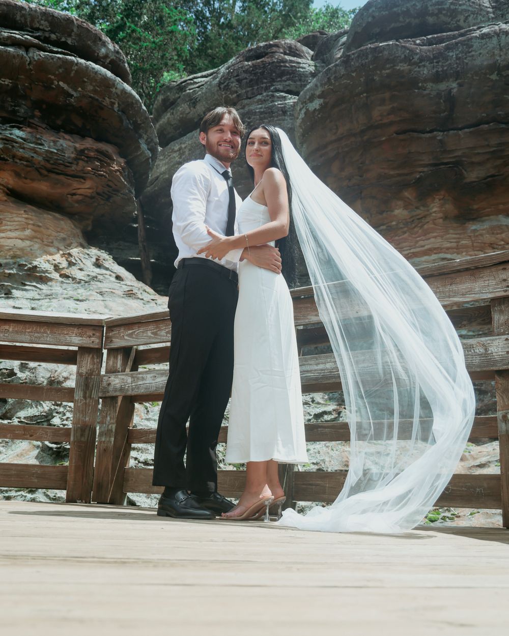 Bride in white dress and veil with groom in black suit posing on wooden deck against sandstone rocks.