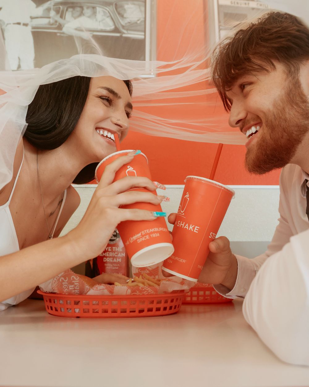 Bride and groom in wedding attire sharing milkshakes at a fast food restaurant, smiling and clinking orange cups.