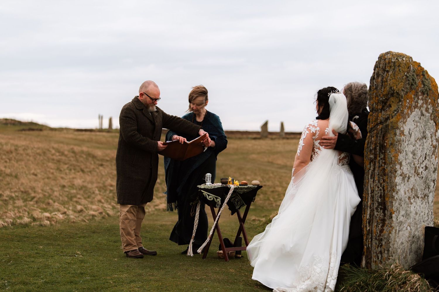 An outdoor wedding ceremony takes place on a grassy hilltop beside a large standing stone on an overcast day.