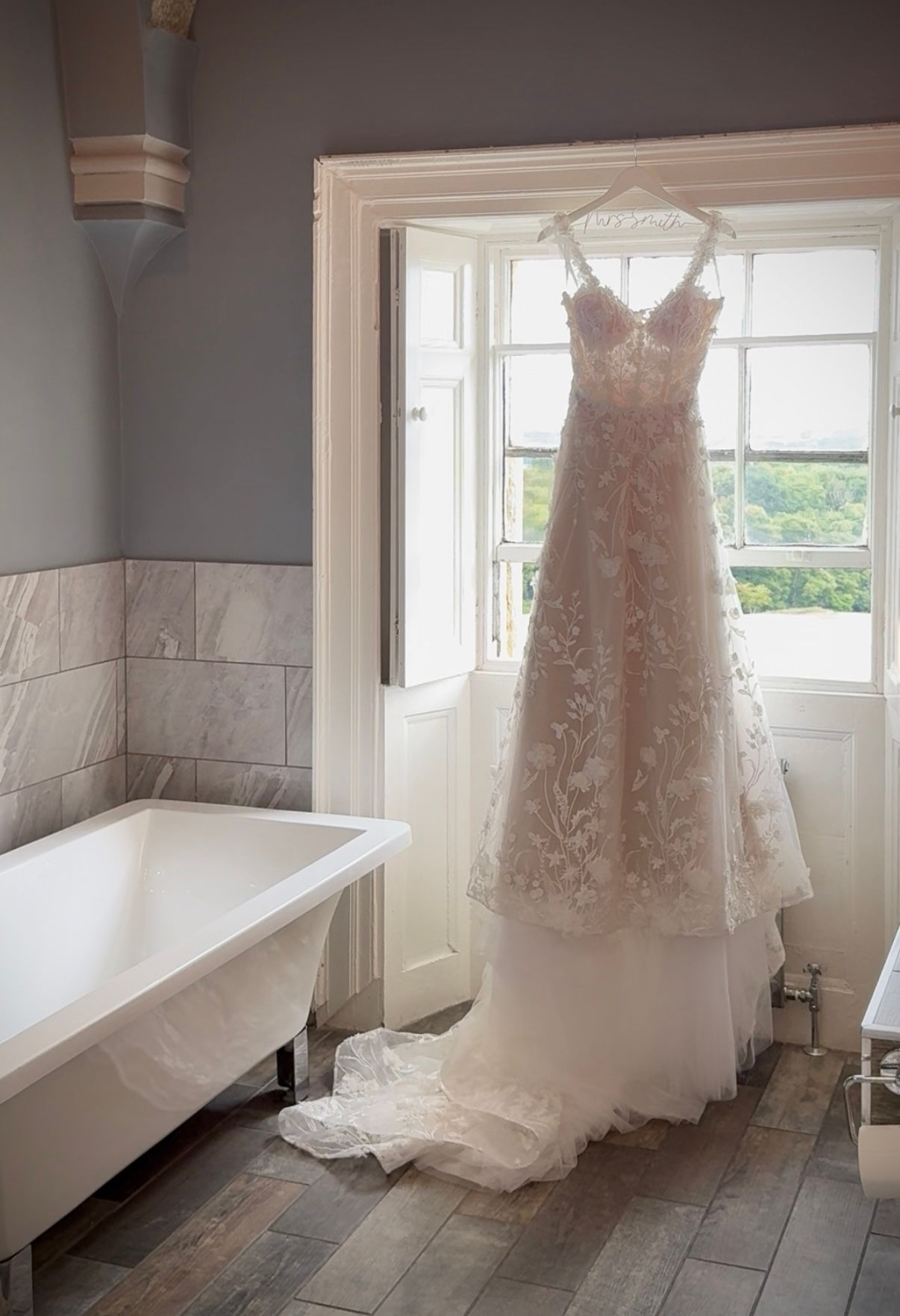 3D lace wedding dress hanging in a tall window of the venue with subtle light shining through the window through the lace bodice.