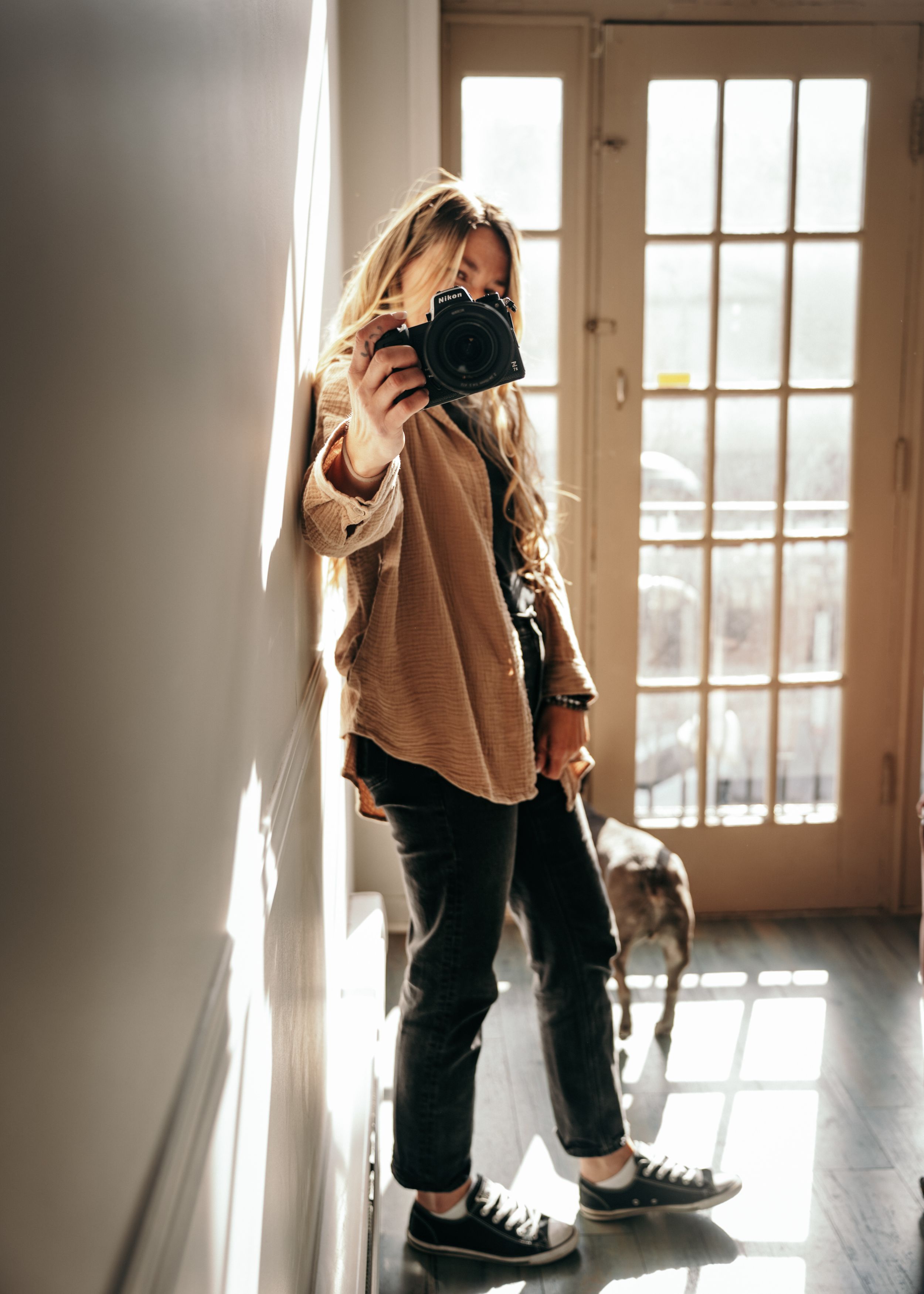 Photographer in tan sweater and jeans takes self-portrait with camera while standing by sunlit French doors.