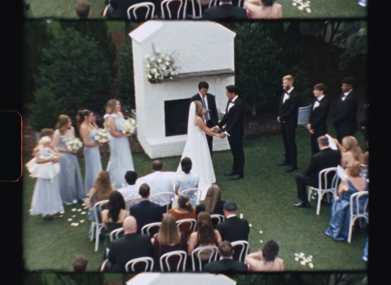 Aerial view of an outdoor wedding ceremony with bridesmaids in white dresses and groomsmen in dark suits gathered on green lawn.