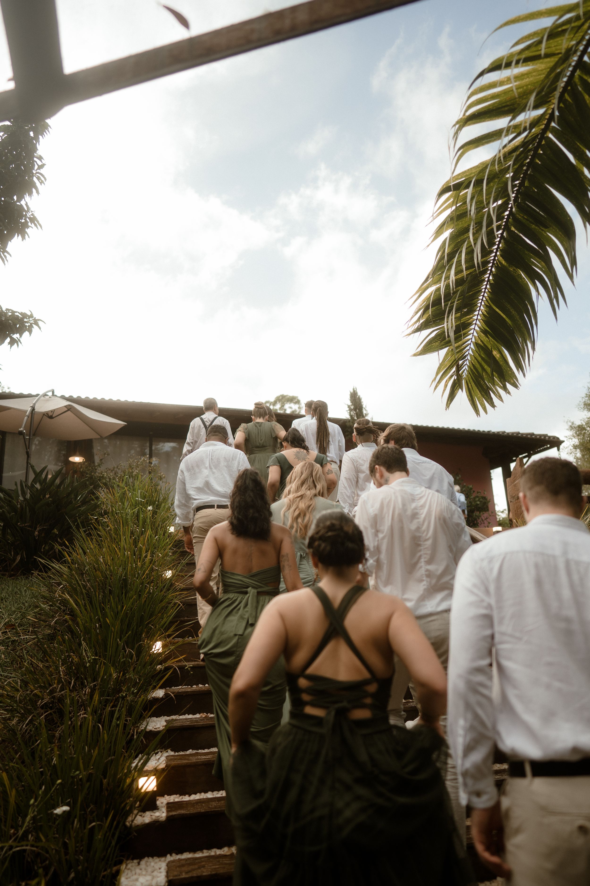 Wedding guests in olive green dresses and white shirts climbing outdoor stone steps under a palm frond.
