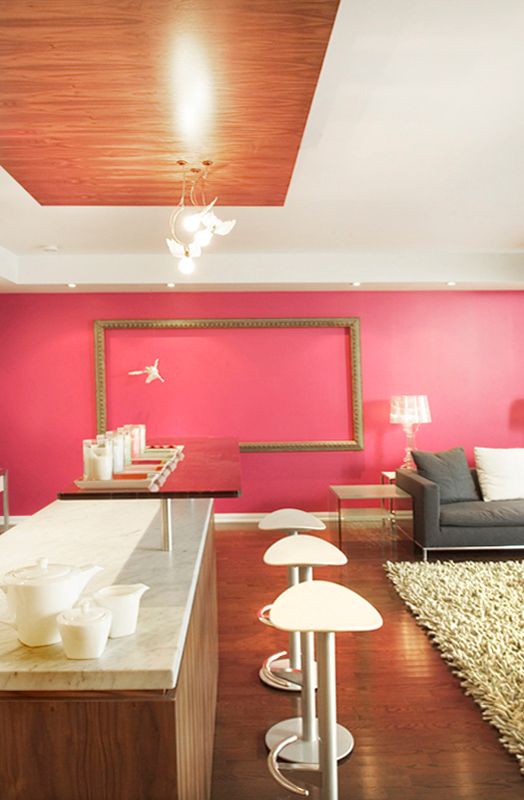 Modern room interior with bright pink accent wall, wooden counter, white bar stools and grey sofa on hardwood floors.