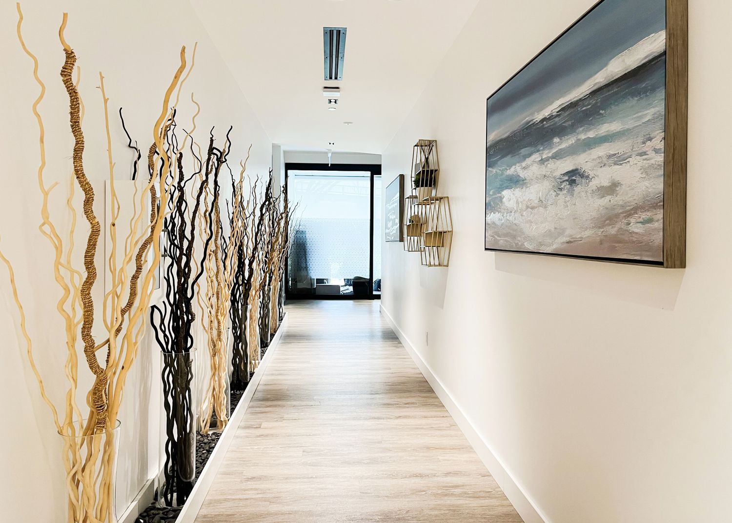 Modern hallway with light wood flooring, decorative branches, and a coastal artwork on white walls leading to a window.