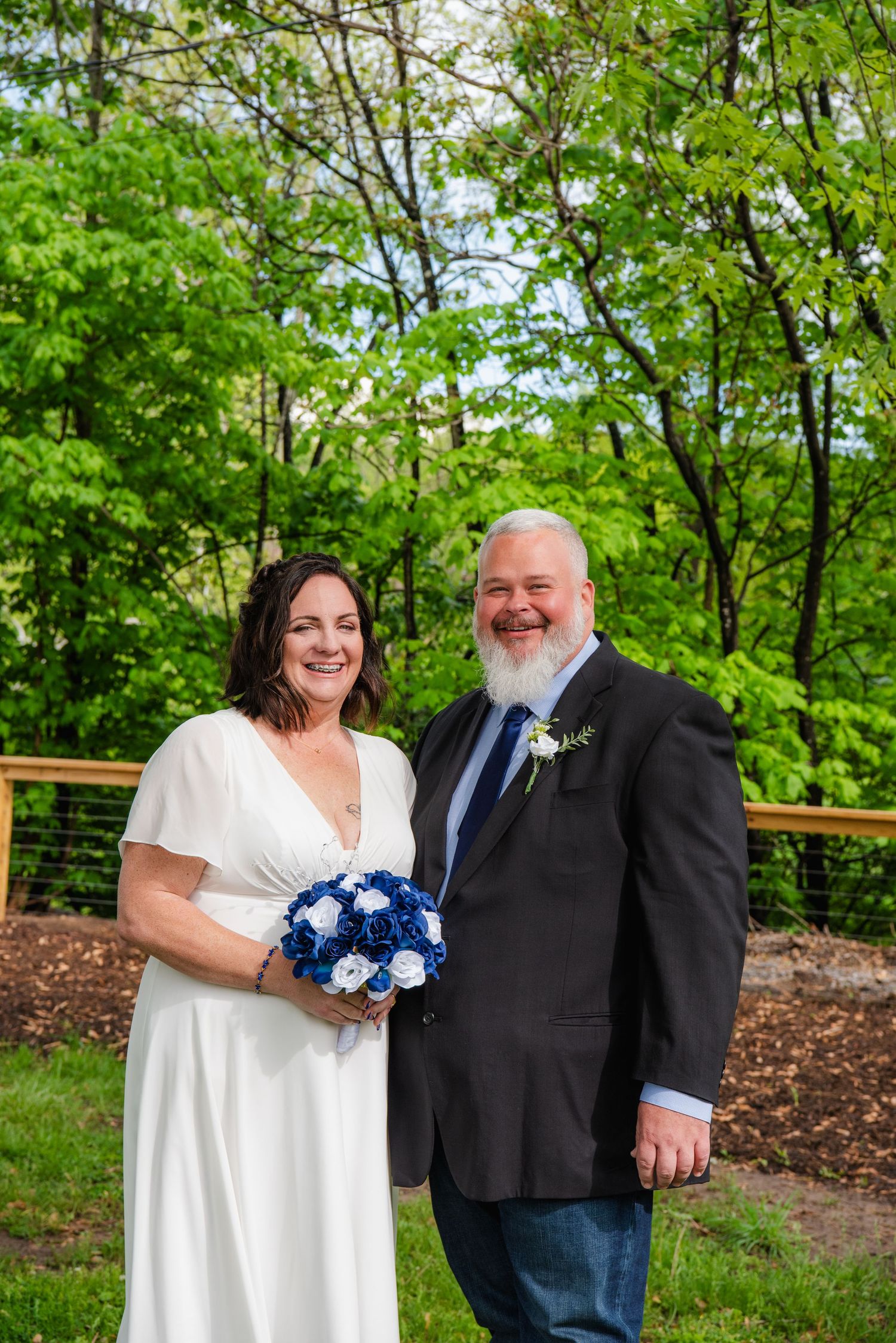 A couple poses outdoors on their wedding day with a blue bouquet against a backdrop of lush green trees.