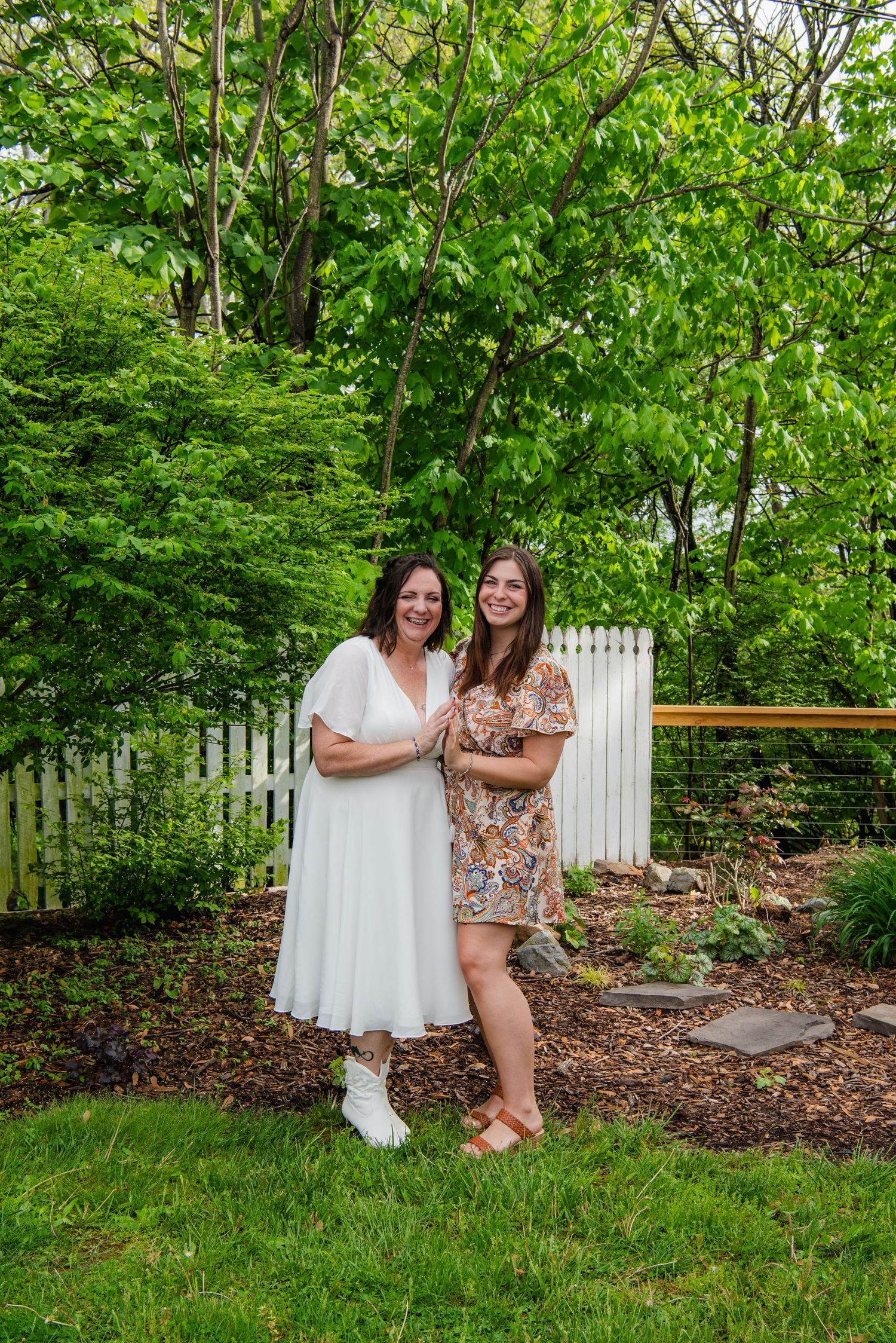 Two women stand together in a garden setting with a white picket fence in the background.