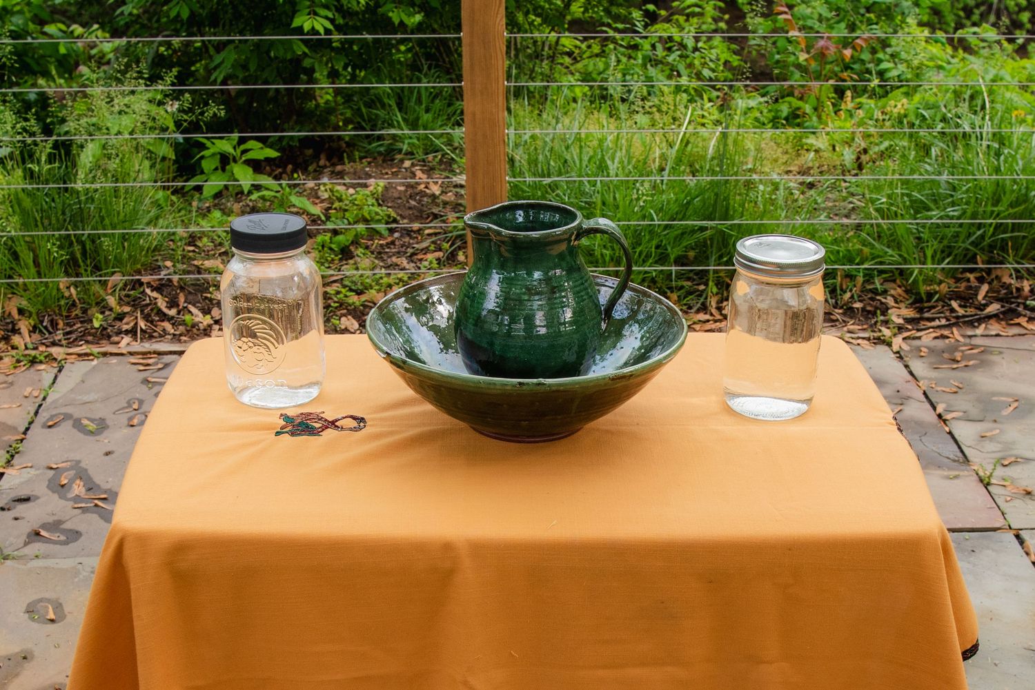 A rustic outdoor ceremony setup with mason jars and a green teapot on an orange tablecloth.