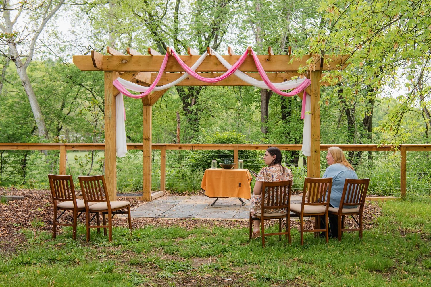A wooden pergola decorated with pink fabric stands in a wooded area with chairs arranged for a ceremony.