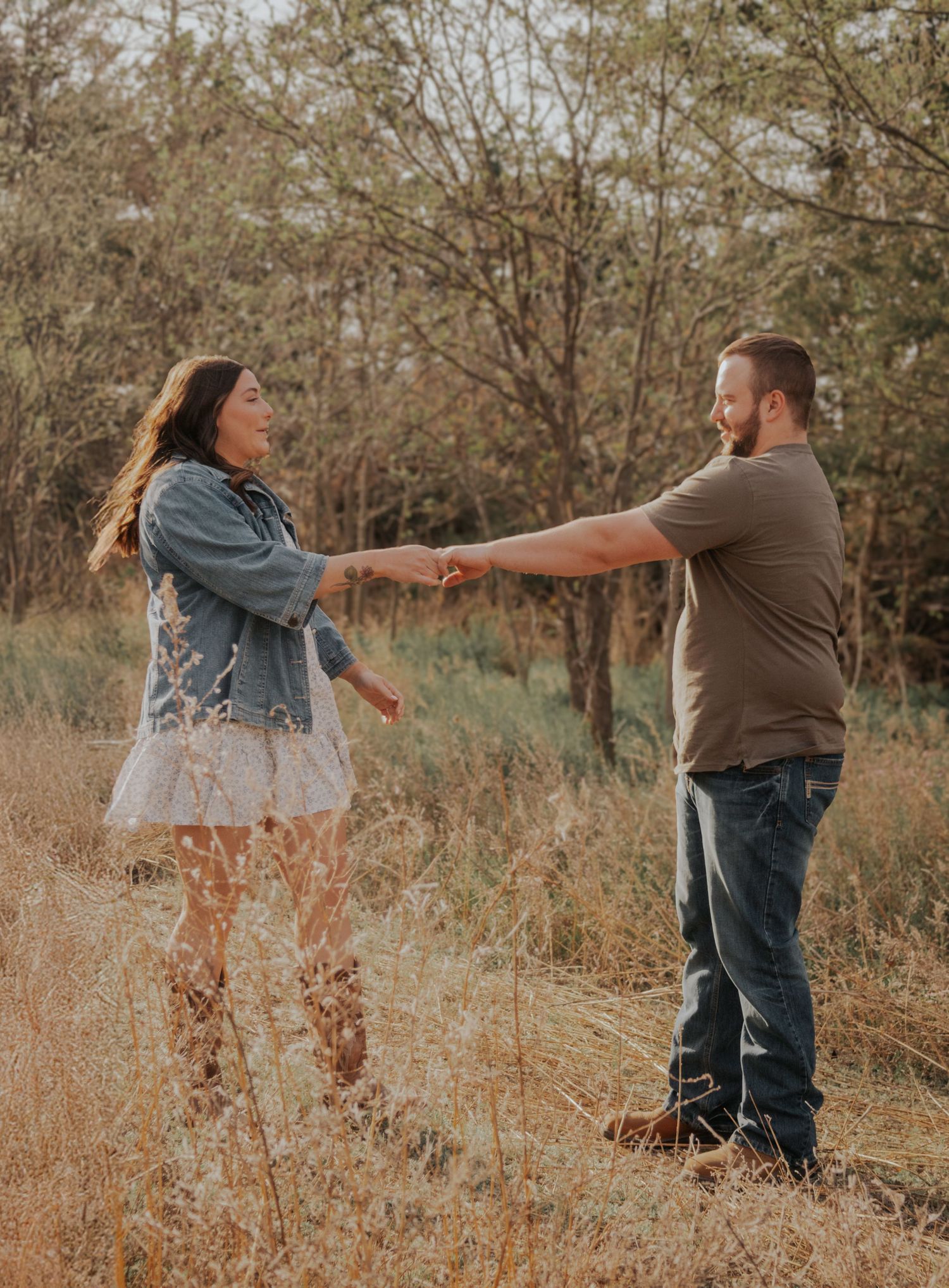 A romantic couple holds hands in a golden field during sunset while wearing casual attire.
