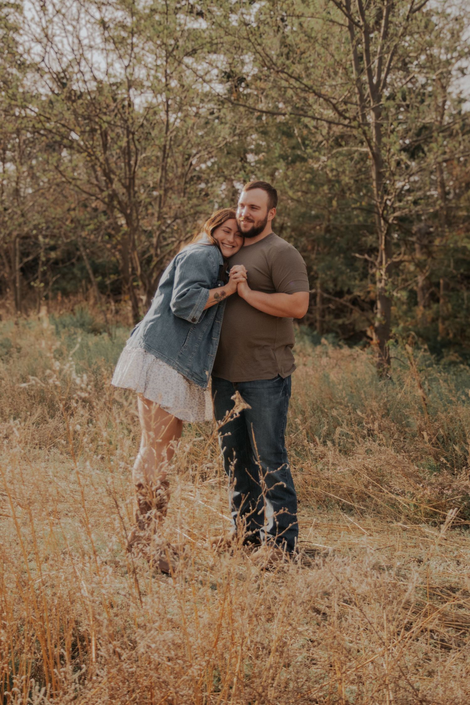A couple embraces and laughs together in a golden field with autumn trees in the background.