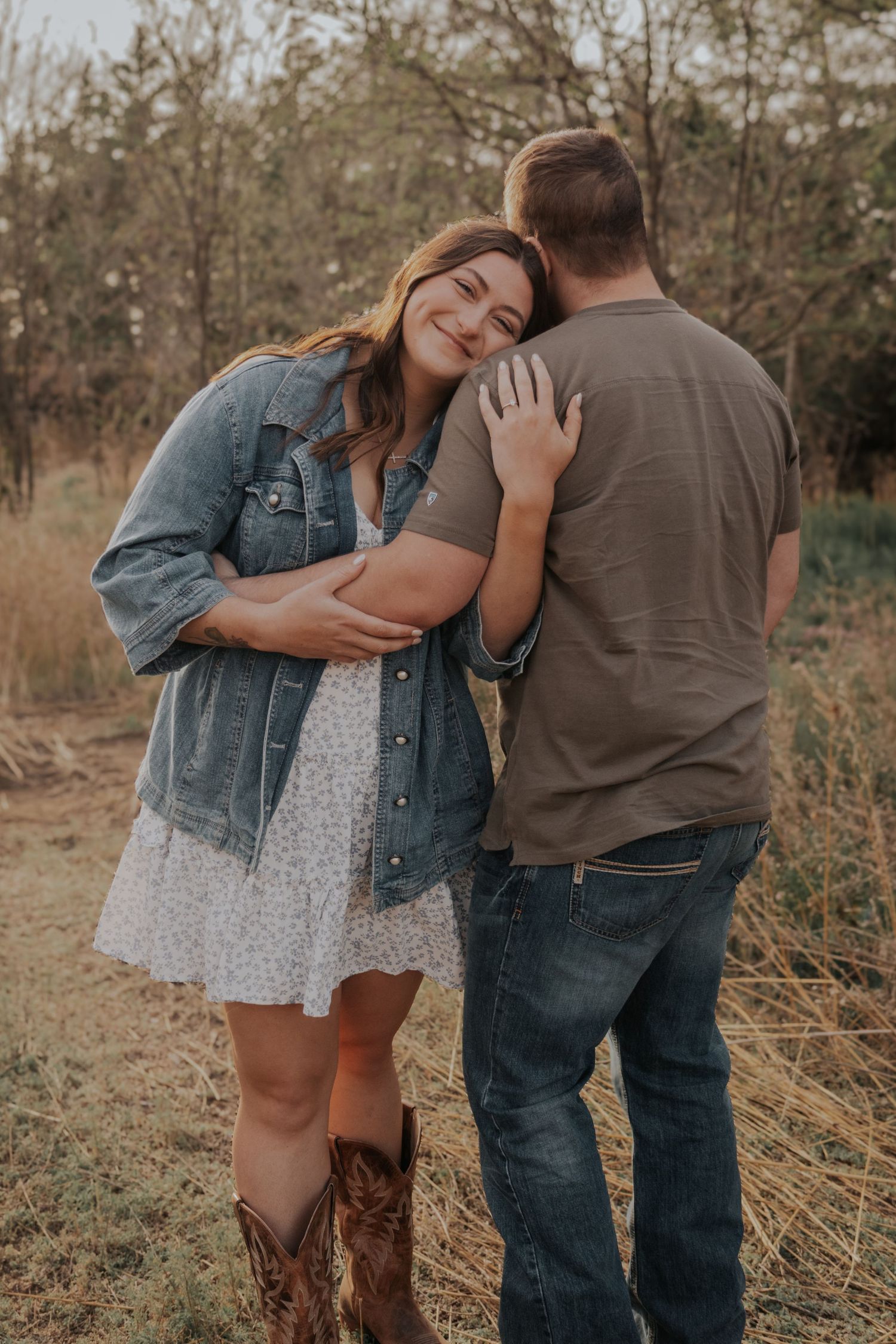 Romantic countryside engagement photo shows couple in casual denim and cowboy boots embracing in a rustic field setting.