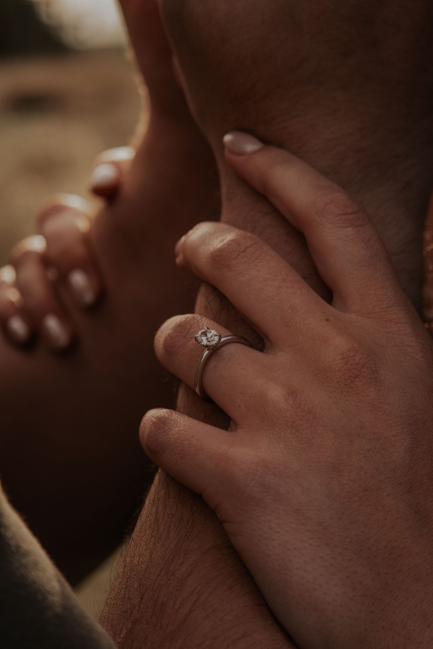 Close-up shot of hands with an engagement ring resting against each other in warm lighting.