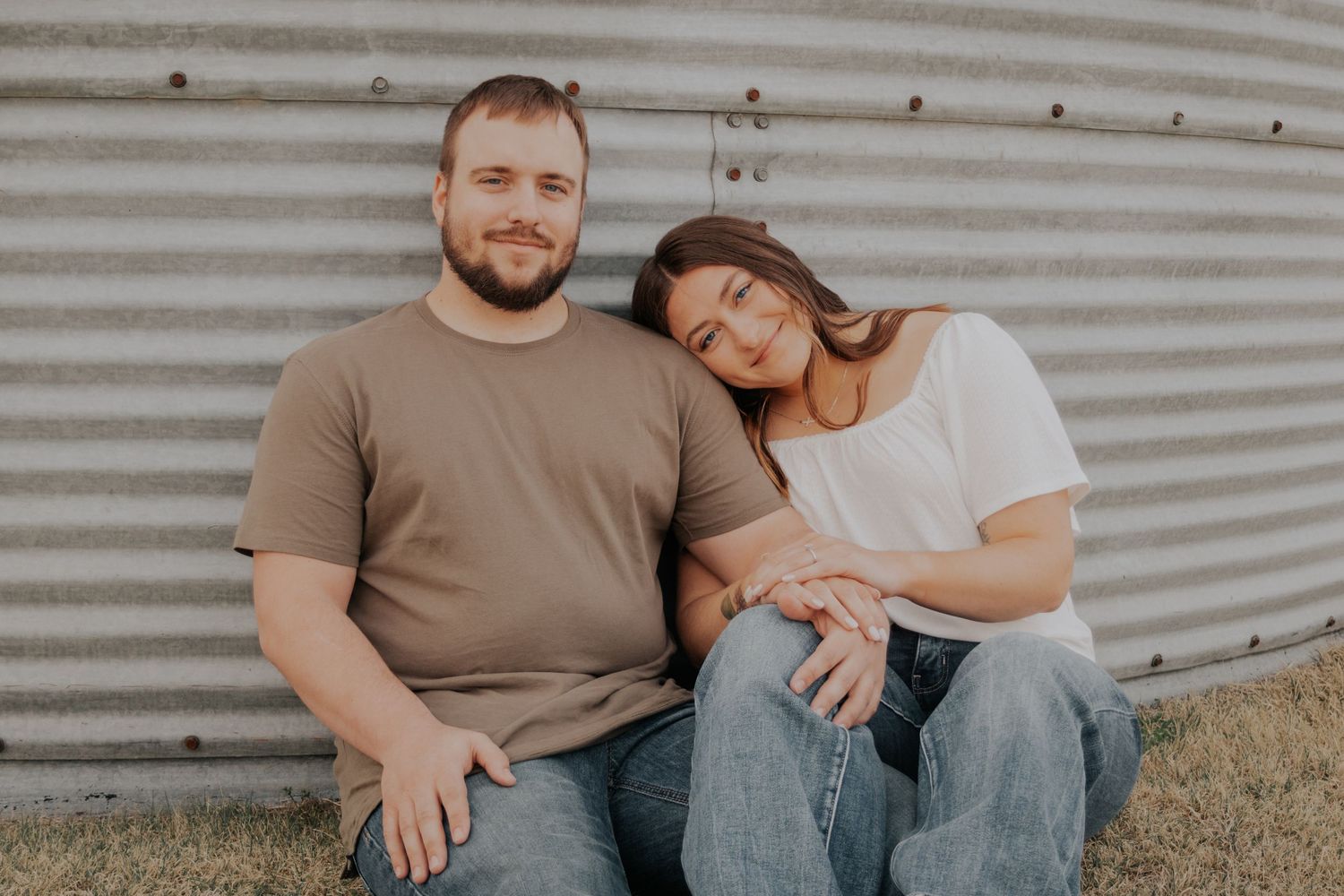 A couple sits close together against a metal wall in soft, natural lighting.