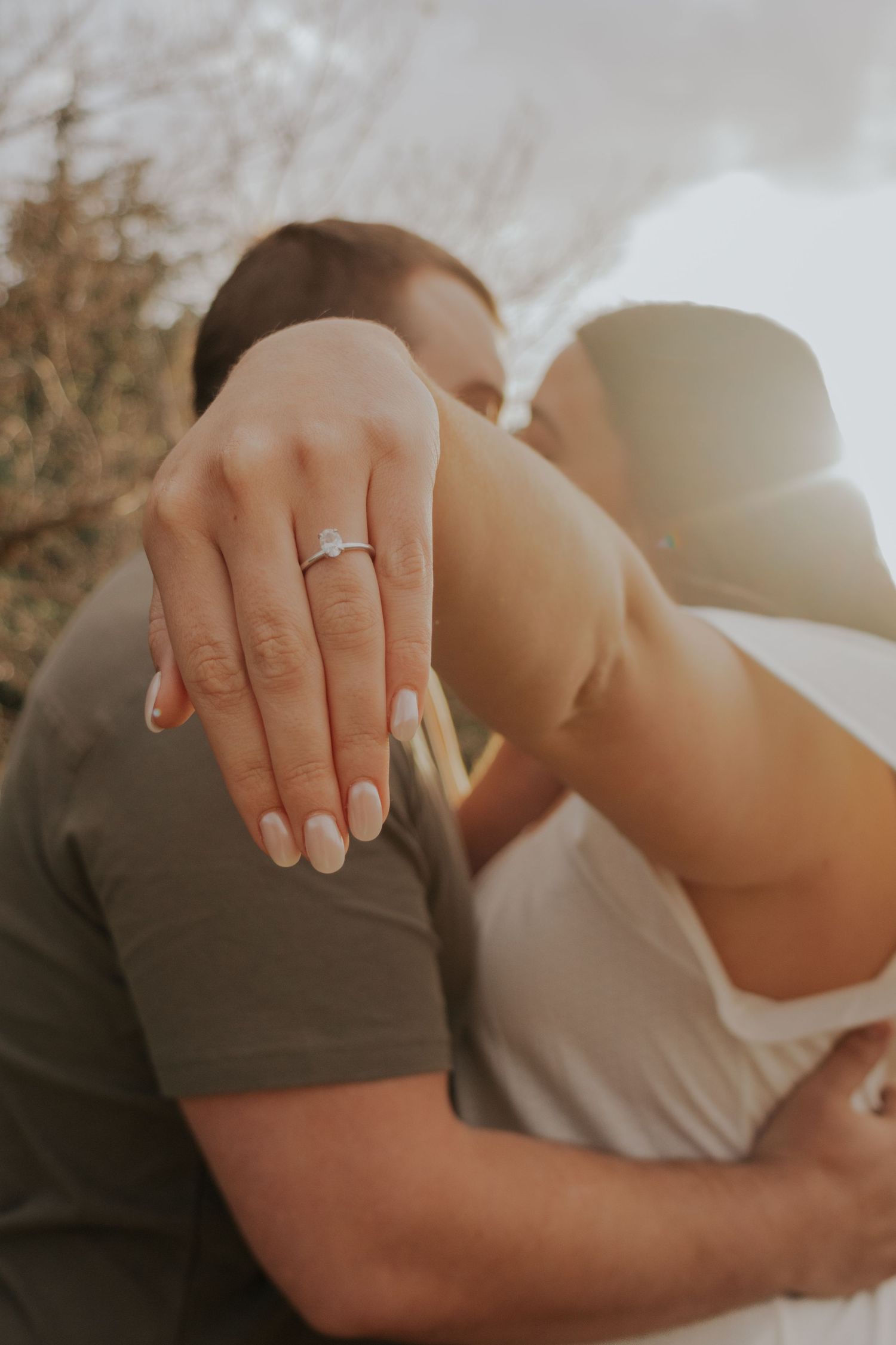 Engaged couple embraces during sunset as woman shows off engagement ring on finger in romantic outdoor photo.