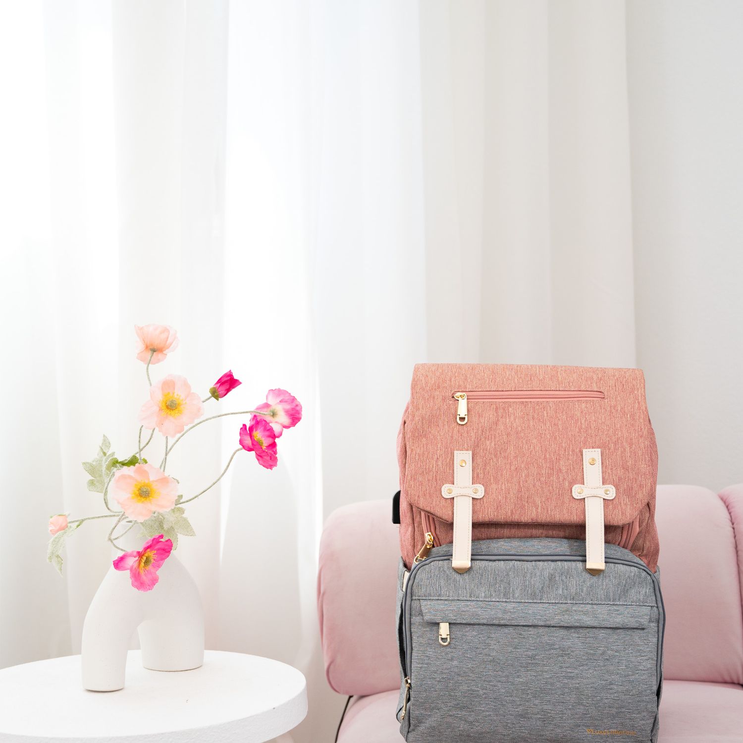 Stylish pink and gray backpack next to delicate pink and coral flowers in white vase on minimal white table setting.