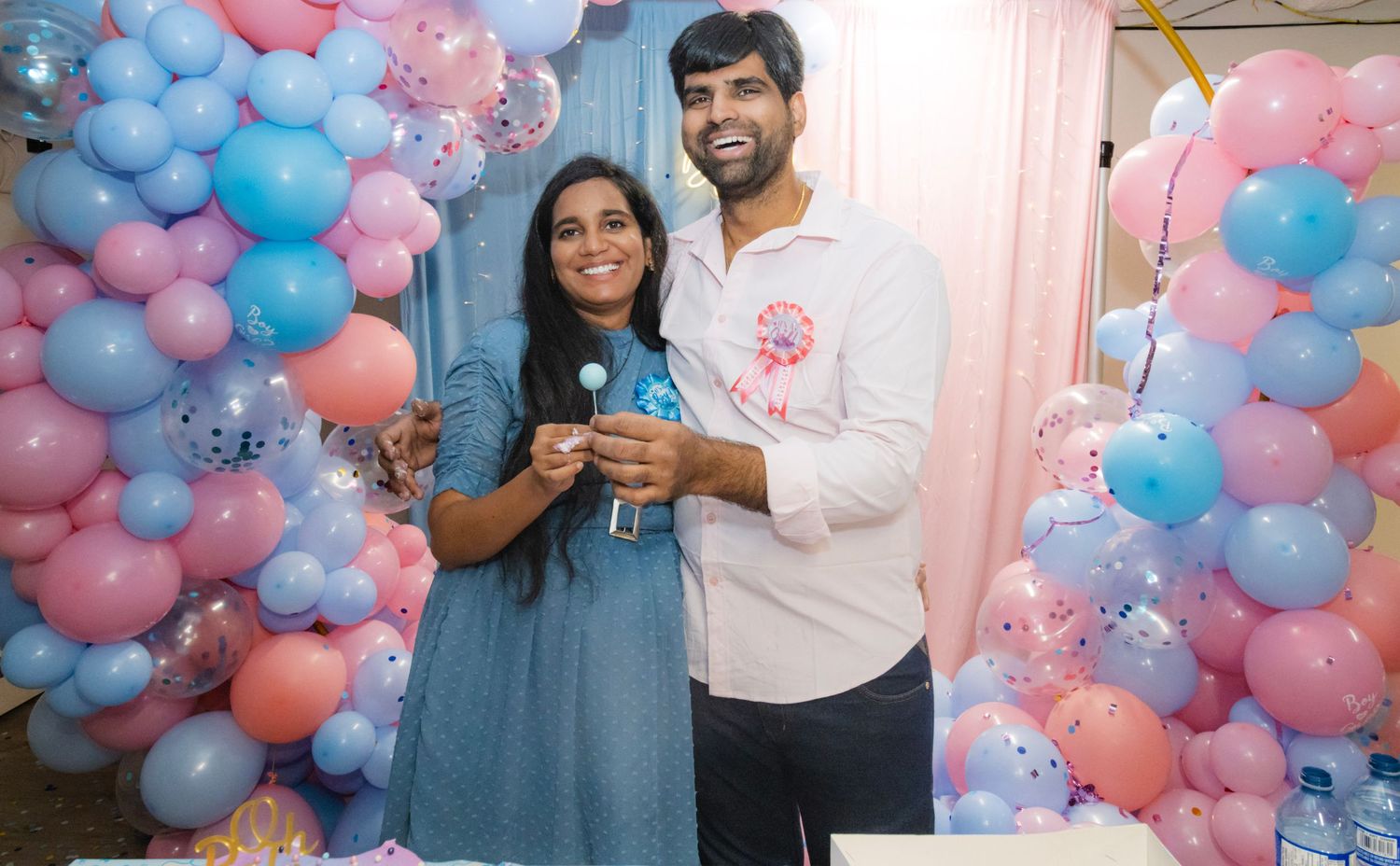 smiling couple at their gender reveal event with blue and pink balloons in the background