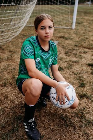 Young soccer player in green jersey sits on grass field by goal net holding white ball.