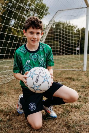 Youth player in green jersey crouches with soccer ball by goal net on dry grass field.