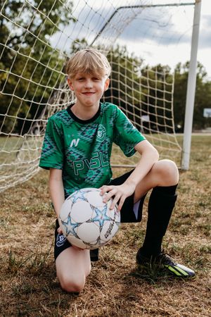 Young soccer player in green jersey kneels with ball in front of goal net during outdoor practice session.