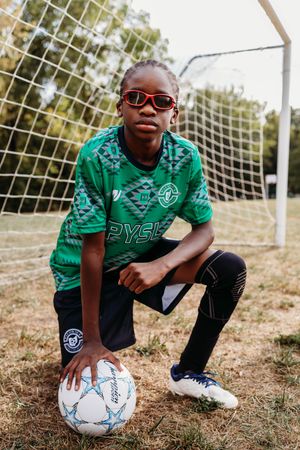 A young soccer player in a green jersey kneels with a soccer ball next to a goal net on a sunny day.