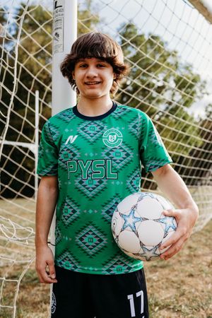 A young soccer player in a green PYSL jersey poses with a white soccer ball in front of goal netting.