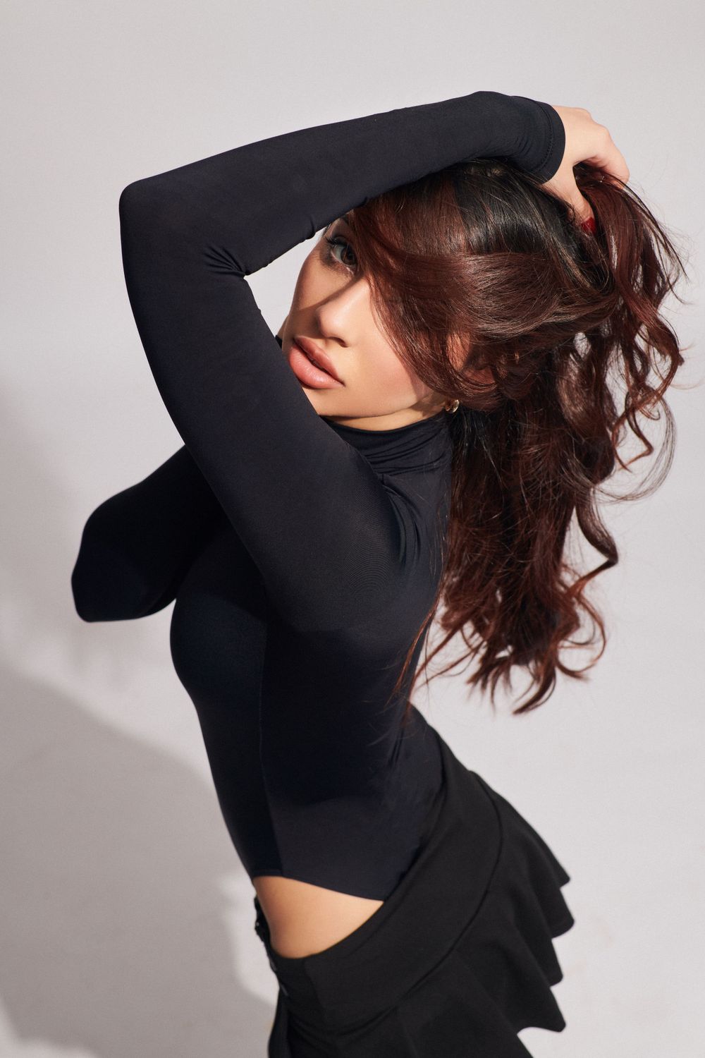 Woman with curly brown hair in a black outfit, posing with one hand on her head against a light gray background.