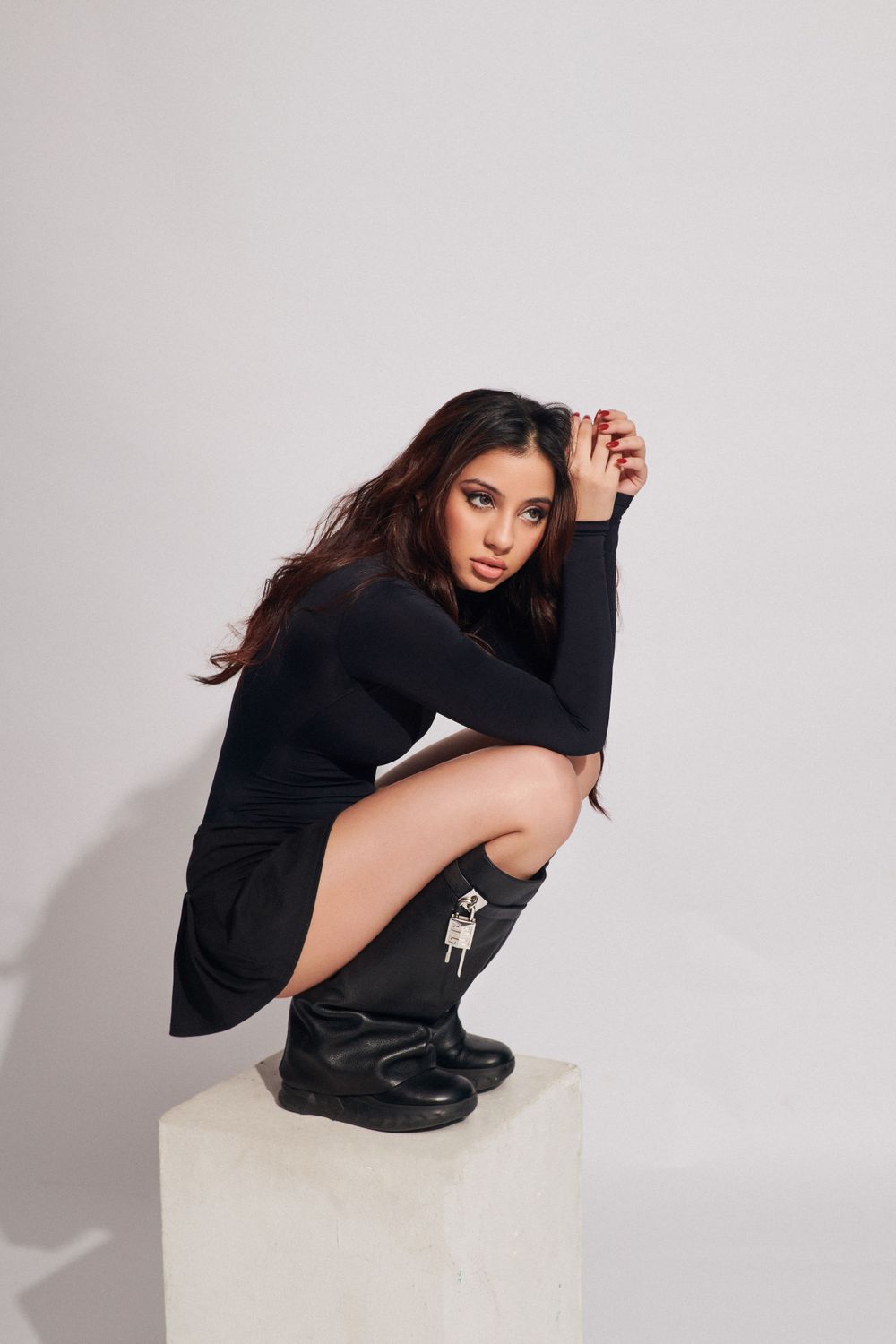Woman in black outfit kneeling on the floor, hand on knee, striking a confident pose against a light gray background.