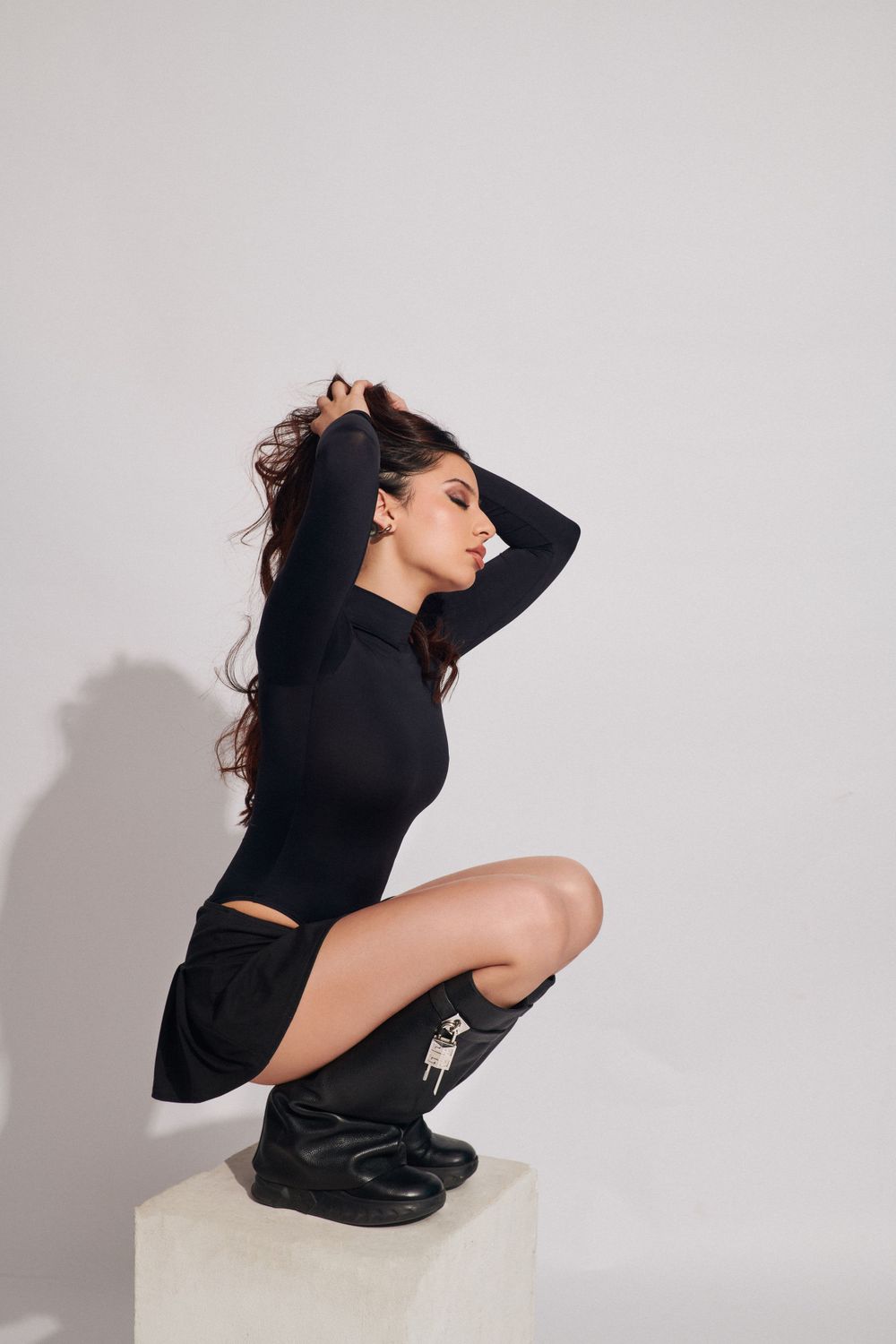 Woman in black outfit kneeling on the floor, hand on knee, striking a confident pose against a light gray background.