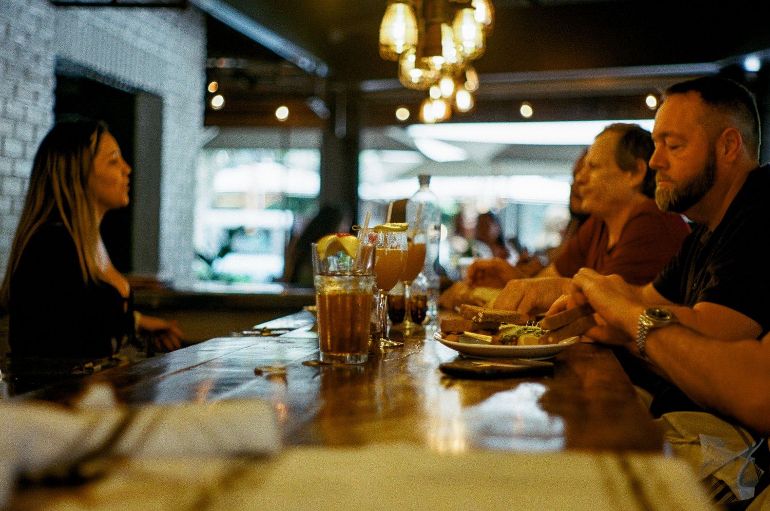 People dining together at a restaurant bar counter with drinks and food in warm ambient lighting.