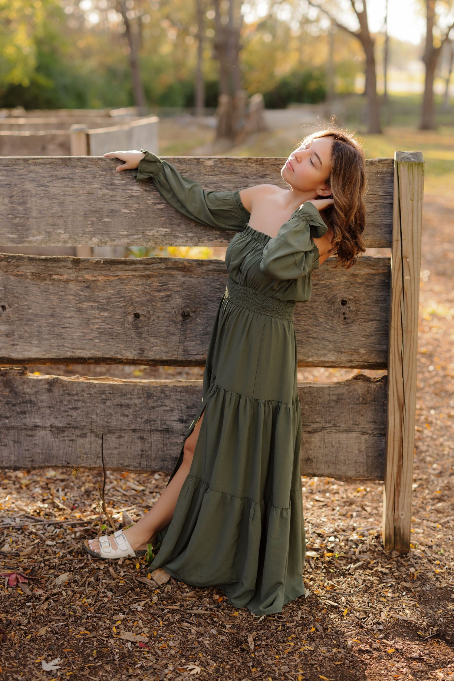 A flowing olive green maxi dress with long sleeves and tiered skirt design photographed against rustic wooden fence in autumn light.