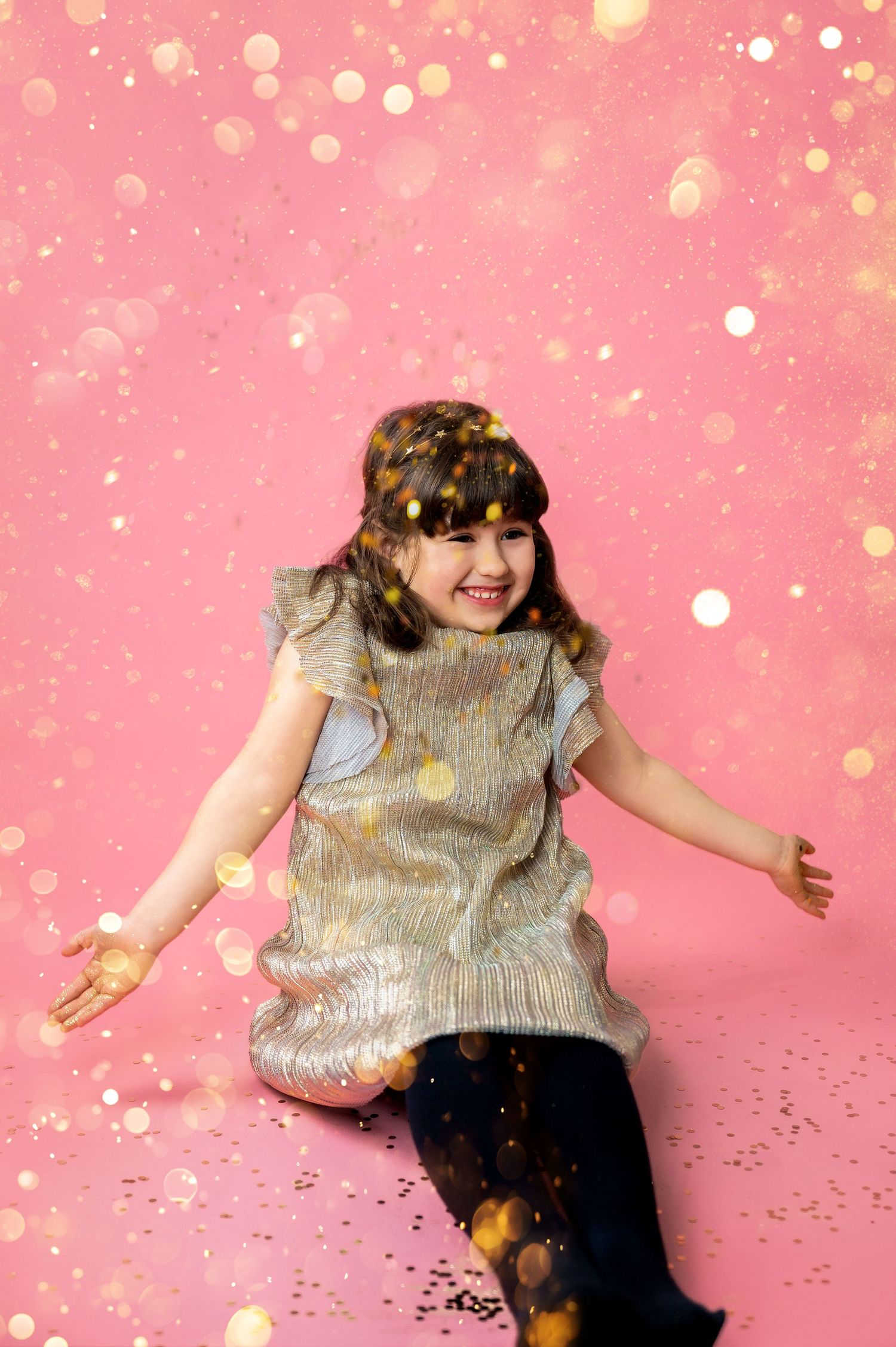 A child wearing a sparkly silver dress and tights sits joyfully against a pink background with glittering bokeh lights.