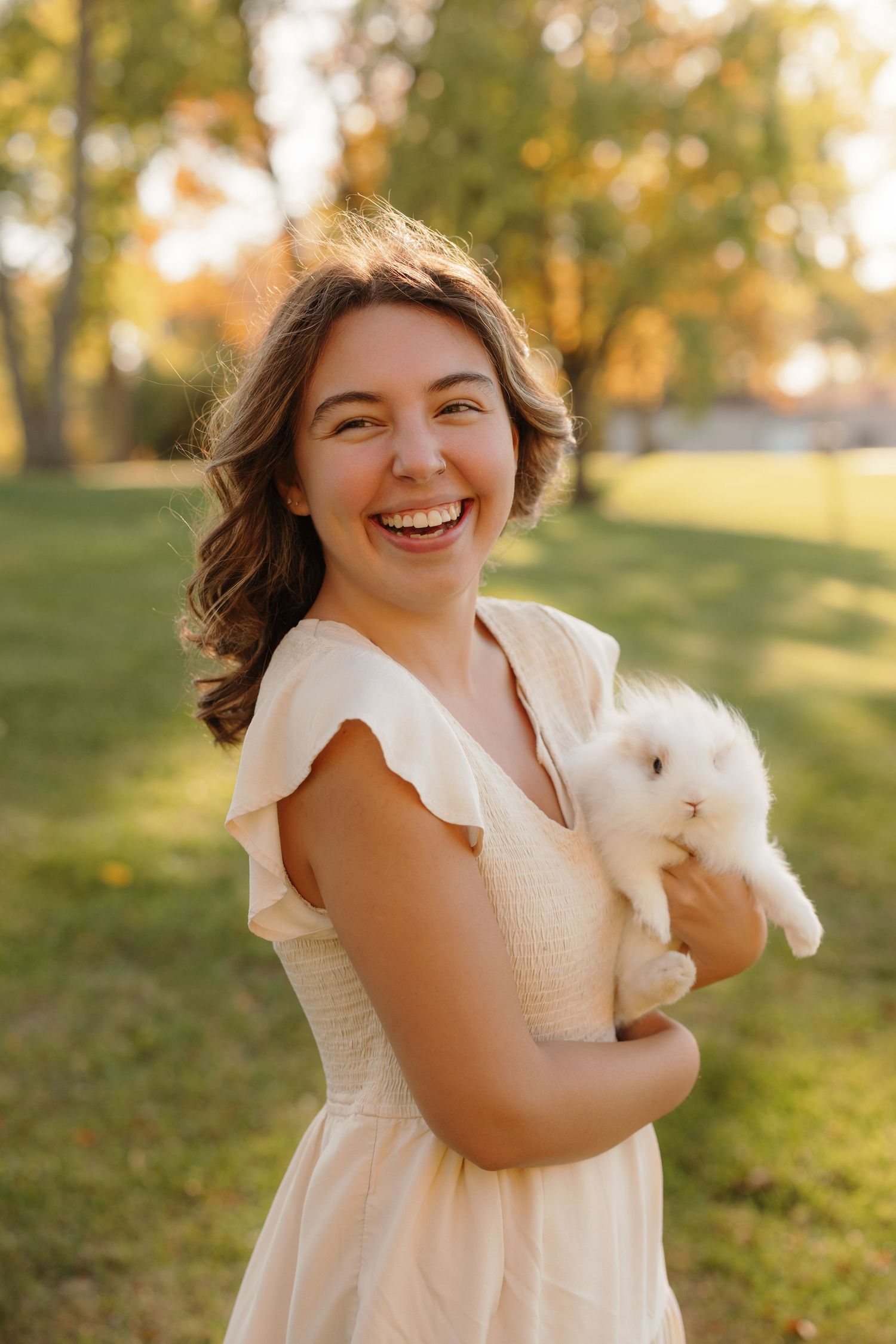 Someone in a white dress laughs joyfully while holding a fluffy white bunny rabbit in an autumn garden setting.